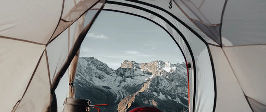 Blick aus einem Zelt auf schneebedeckte Berge, mit einem roten Schlafsack im Vordergrund, sinnbildlich für warme Nächte beim Camping in kalten Höhenlagen.