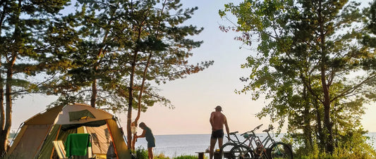 Zeltplatz an einem See bei Sonnenuntergang, mit hohen Baeumen, Fahrraedern und einem aufgestellten Zelt im warmen Abendlicht