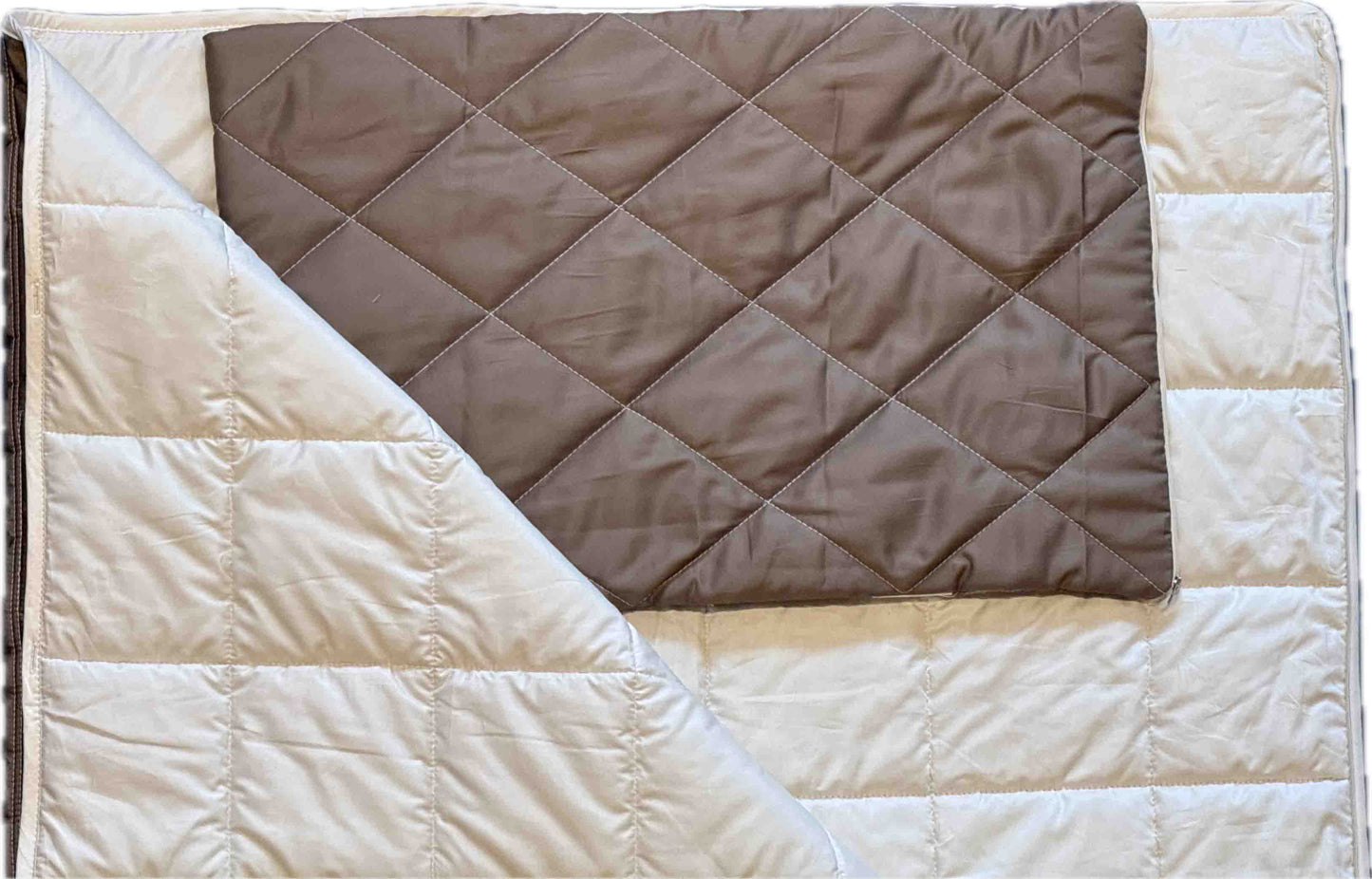 Eine gefaltete braune Kissenhülle „Essential“ liegt auf einer größeren, wärmeisolierenden beigen Steppdecke, deren eine Ecke umgeschlagen ist, sodass das Innenfutter sichtbar wird. Beide sind mit einem aufgestickten Rastermuster verziert.