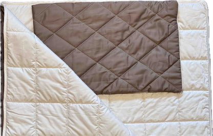 Eine gefaltete braune Kissenhülle „Essential“ liegt auf einer größeren, wärmeisolierenden beigen Steppdecke, deren eine Ecke umgeschlagen ist, sodass das Innenfutter sichtbar wird. Beide sind mit einem aufgestickten Rastermuster verziert.
