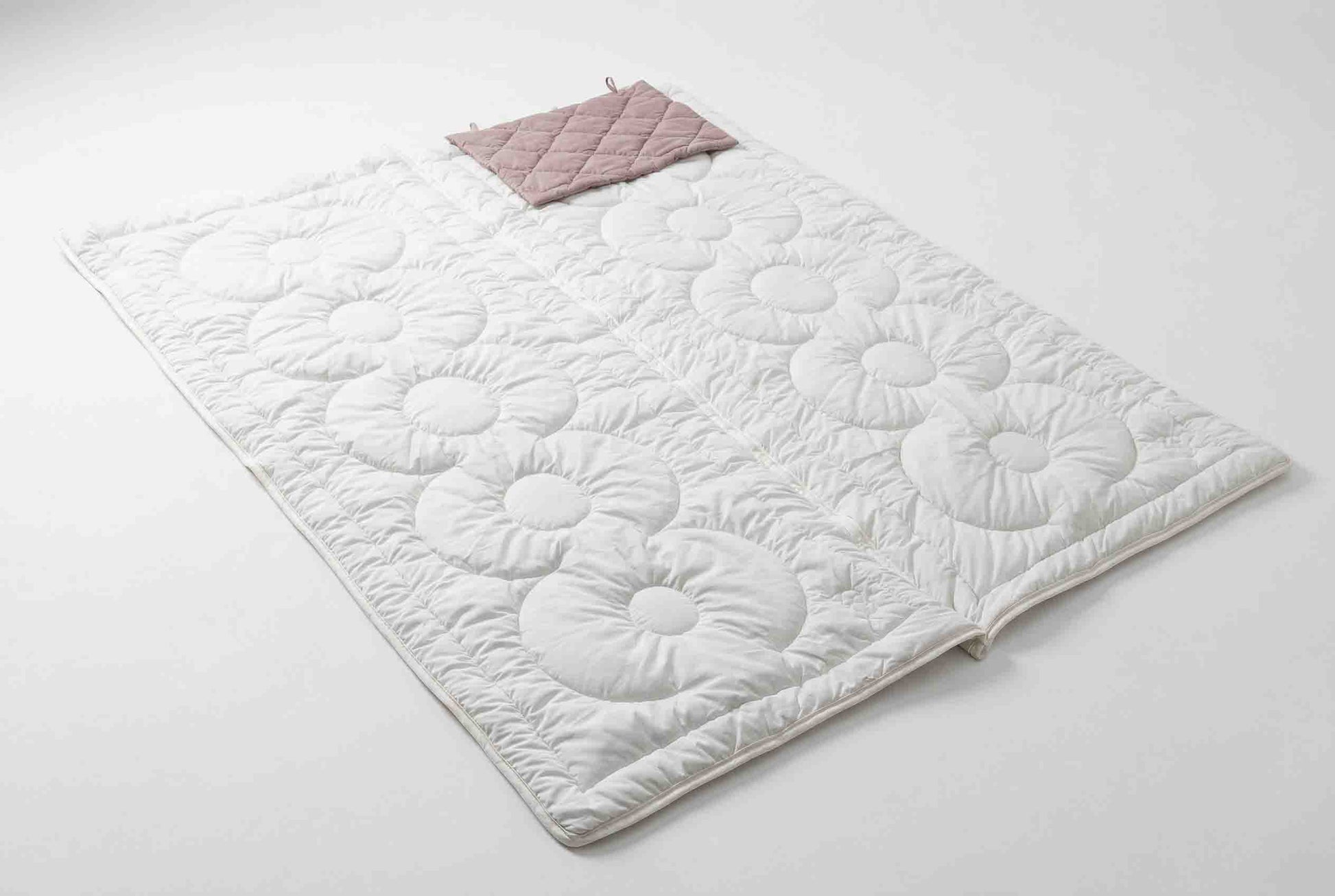 Eine weiße, gesteppte Matte mit Blumenmuster liegt flach auf einer weißen Fläche. An einem Ende sorgt ein zusammengefalteter, hellrosa „Origobag Essential Kamelhaar Schlafsack“ aus nachhaltigen Materialien für umweltfreundlichen Komfort.