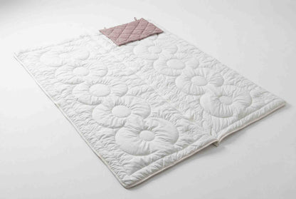 Eine weiße, gesteppte Matte mit Blumenmuster liegt flach auf einer weißen Fläche. An einem Ende sorgt ein zusammengefalteter, hellrosa „Origobag Essential Kamelhaar Schlafsack“ aus nachhaltigen Materialien für umweltfreundlichen Komfort.