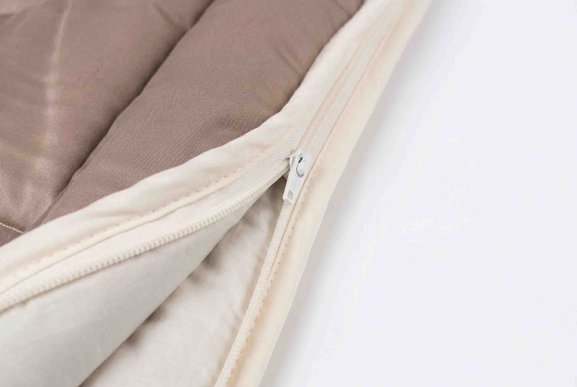 Nahaufnahme der „Origobag Essential Baumwolle“, einer beige-braunen Steppdecke aus nachhaltigen Naturmaterialien, mit einem teilweise geöffneten weißen Reißverschluss, der den Blick auf die Stofflagen und die detaillierten Nähte auf weißem Hintergrund freigibt.