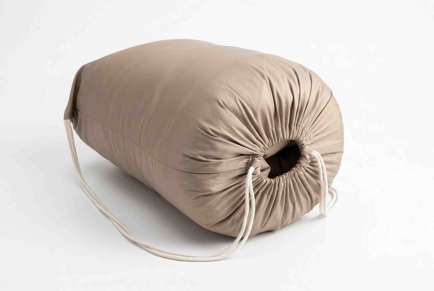 Der „Origobag Premium Baumwolle Seide“, ein zylindrisches, beiges Kissen, handgefertigt und an einem Ende mit einem Kordelzug versehen, liegt auf einer weißen Fläche, wobei die Kordeln locker herabhängen.