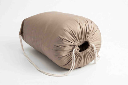 Der „Origobag Premium Baumwolle Seide“, ein zylindrisches, beiges Kissen, handgefertigt und an einem Ende mit einem Kordelzug versehen, liegt auf einer weißen Fläche, wobei die Kordeln locker herabhängen.